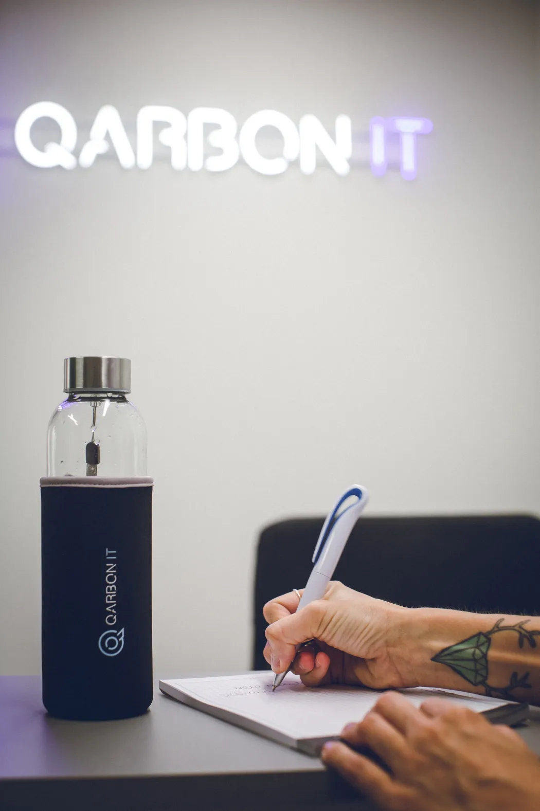 hand taking notes, water bottle and neon sign with qarbon it logo in the background