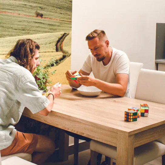 two men sit at a table, eat and solve rubik's cubes